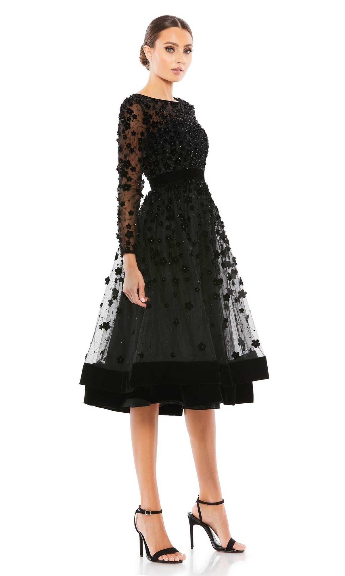 A woman stands against a white background wearing the Mac Duggal 67007 knee-length wedding-guest dress, featuring floral embellishments, black hue, long sheer sleeves, and paired with black high-heeled sandals.